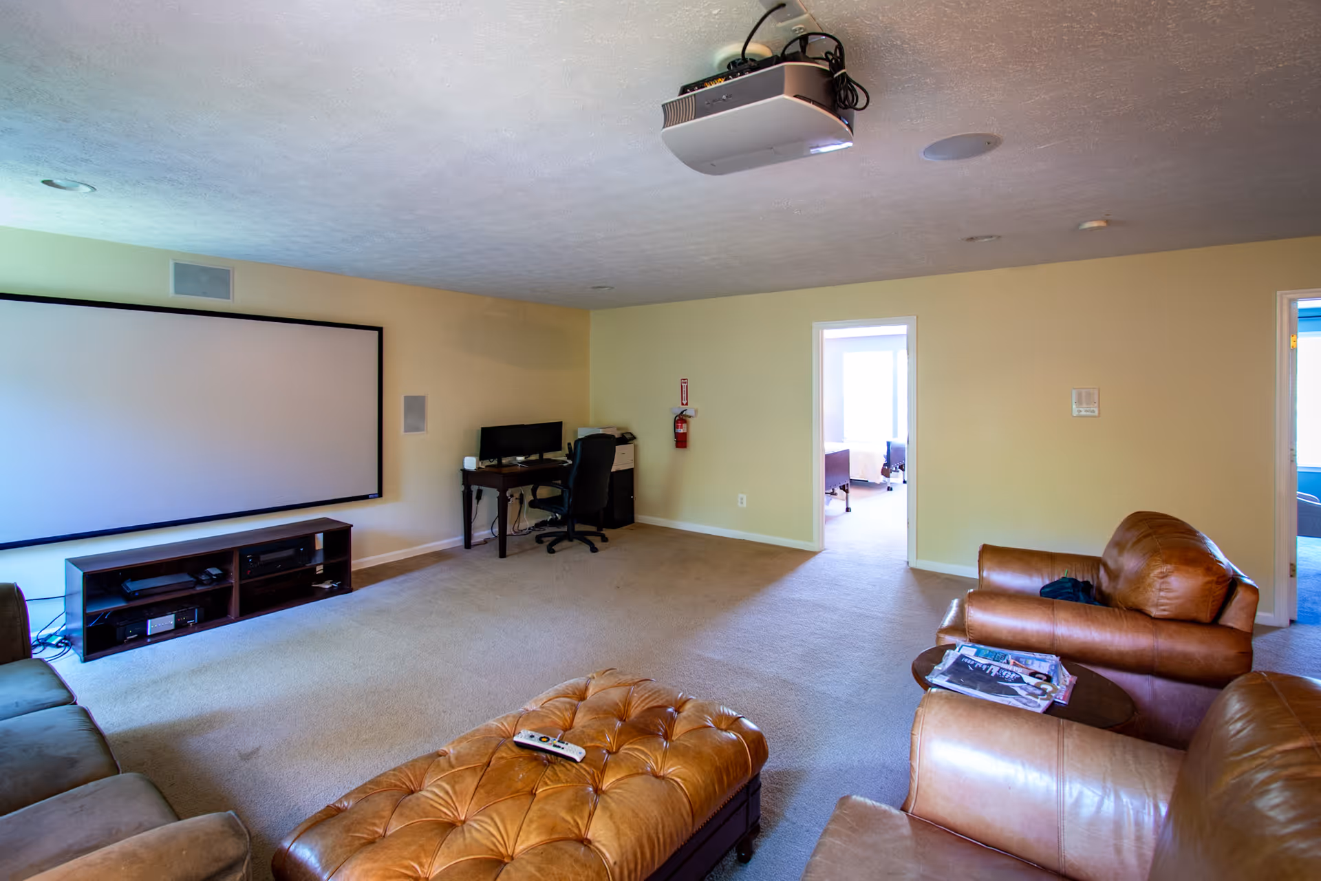 A spacious living room with beige walls and carpeted floor, featuring a large projector screen on one wall, a brown leather sofa and armchairs, a tufted leather ottoman with a remote control on it, a small wooden table with magazines, and a desk with a computer and office chair in the corner. Two doorways lead to other rooms with visible furniture.