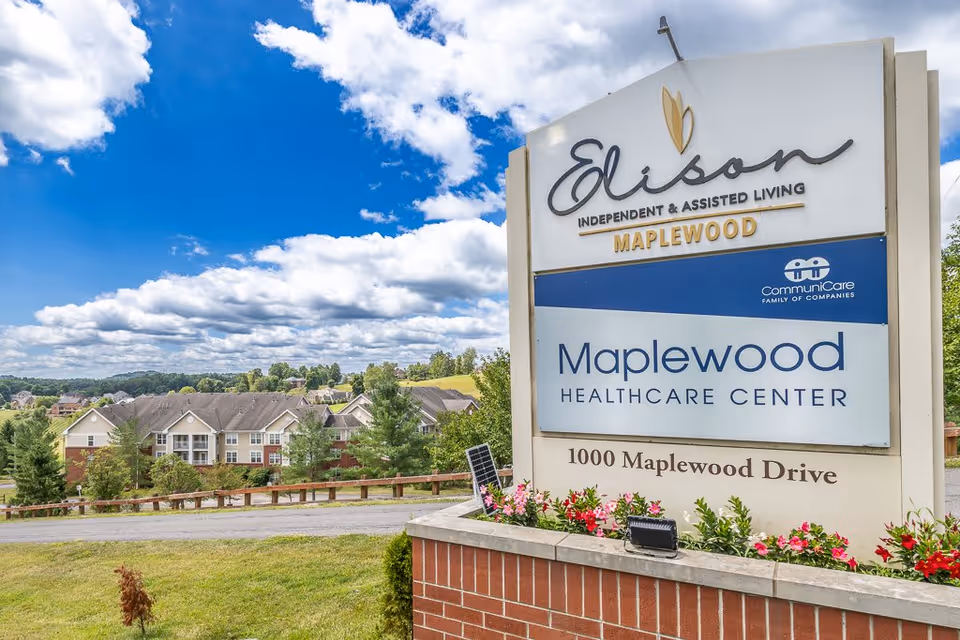 Outdoor view of the Elison Independent & Assisted Living of Maplewood sign with a partly cloudy blue sky and residential buildings in the background.