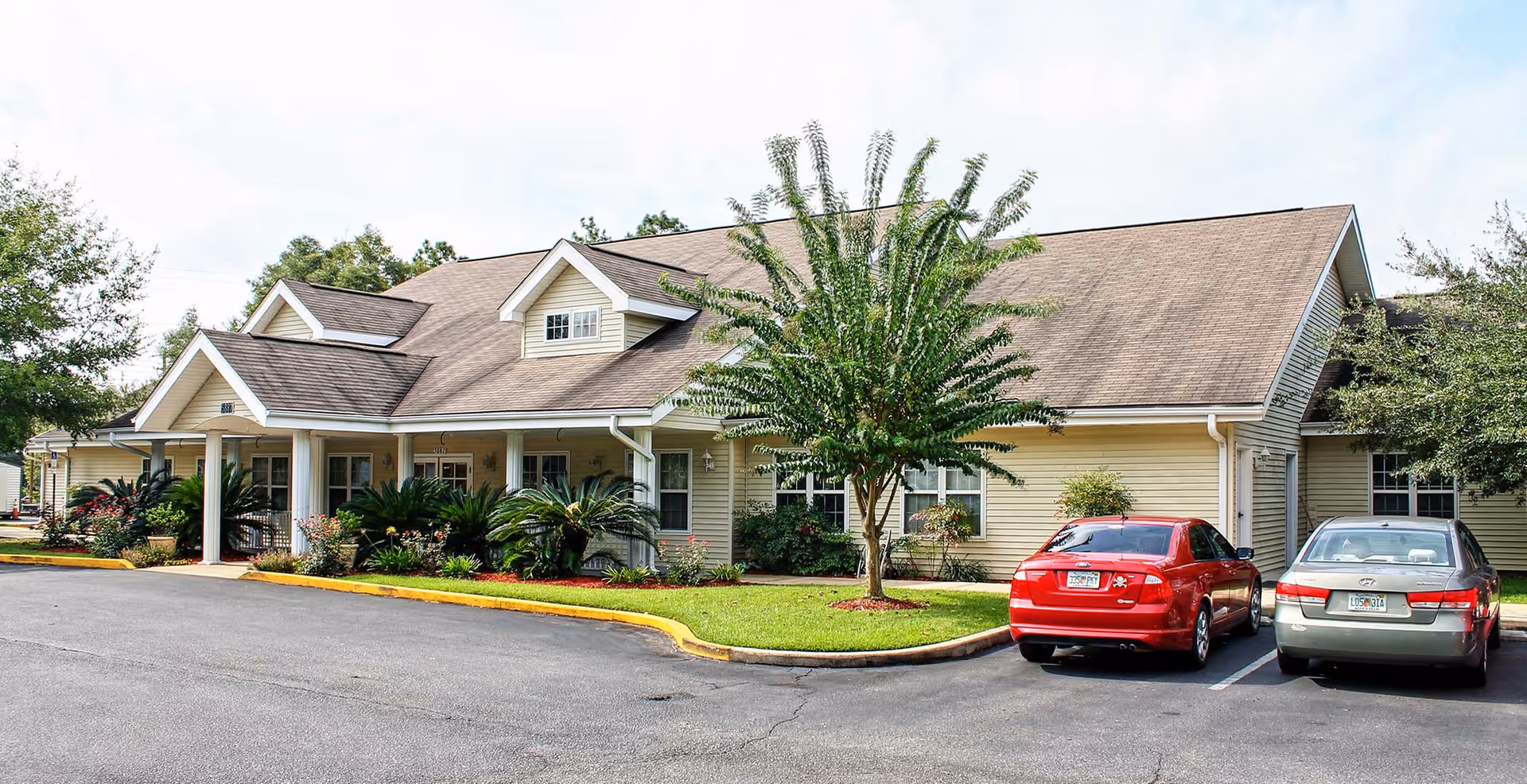 Exterior view of a single-story building with beige siding and a brown shingled roof, surrounded by greenery and two parked cars in front.