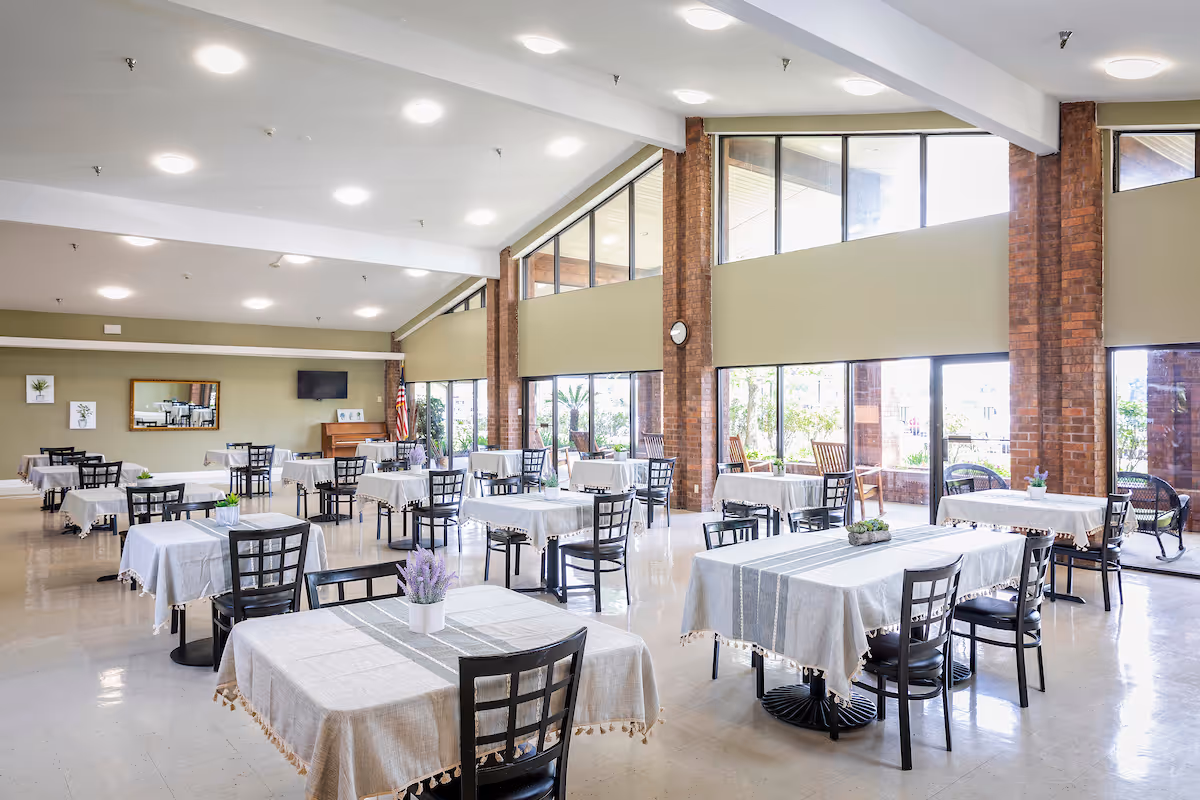 A spacious dining room with multiple tables covered in light-colored tablecloths, each surrounded by black chairs. The room has large windows letting in natural light, brick pillars, and a piano with an American flag in the corner. Small plants and flowers decorate the tables.