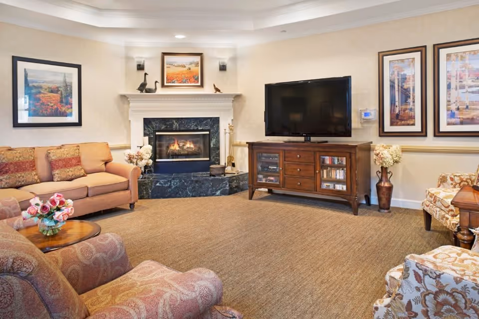 A cozy living room with a beige sofa and patterned armchairs surrounding a small wooden coffee table with a vase of pink roses. There is a fireplace with a marble surround and a mantel decorated with small sculptures and a framed painting above it. A large flat-screen TV sits on a wooden cabinet with glass doors. The walls are adorned with framed landscape artwork, and the floor is carpeted in a neutral tone.