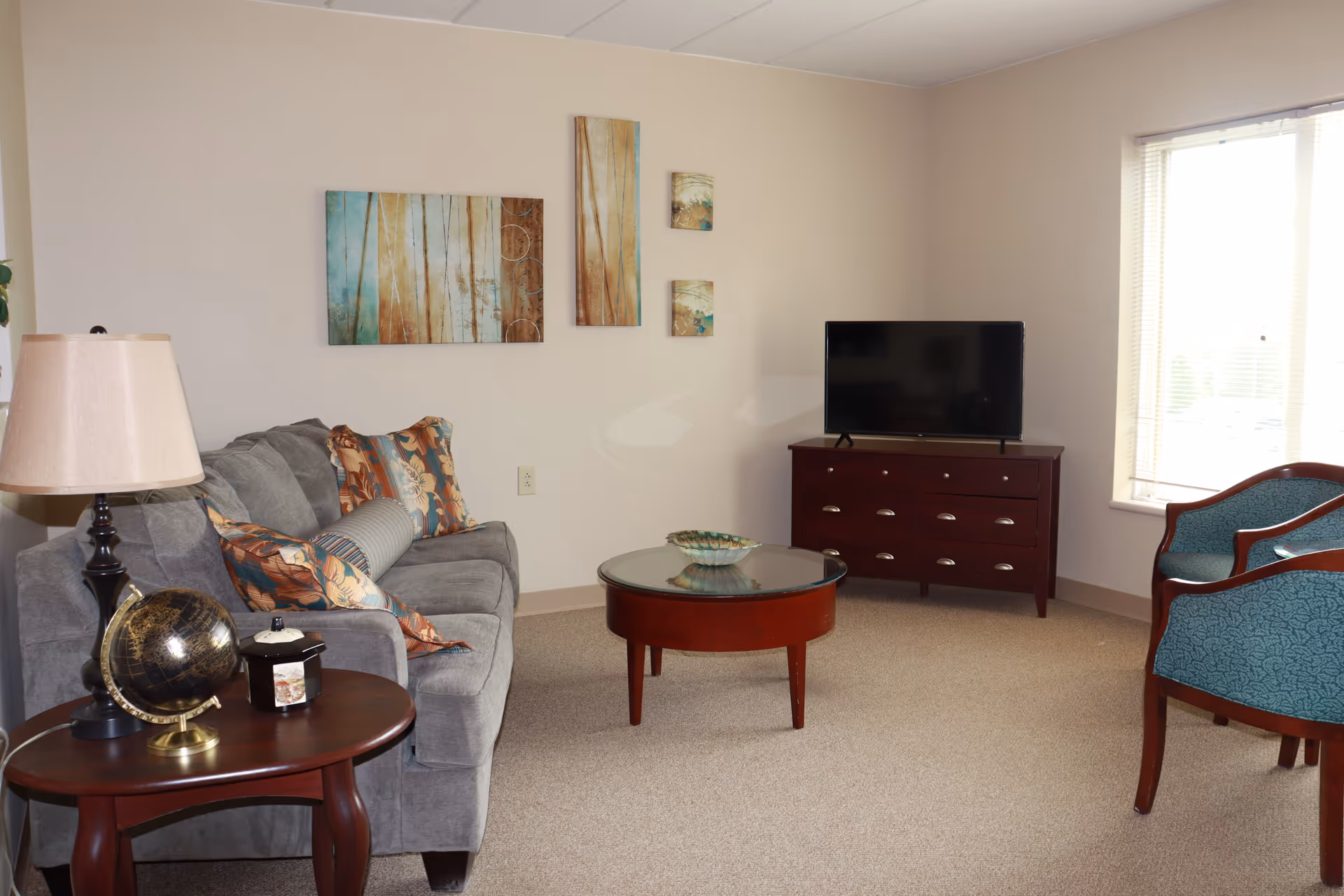 A cozy living room with a gray sofa adorned with patterned cushions, a round wooden coffee table with a glass top, a wooden side table with a lamp and a decorative globe, two teal upholstered chairs, a wooden TV stand with a flat-screen television, and abstract wall art on a beige wall. A window with blinds allows natural light into the room.