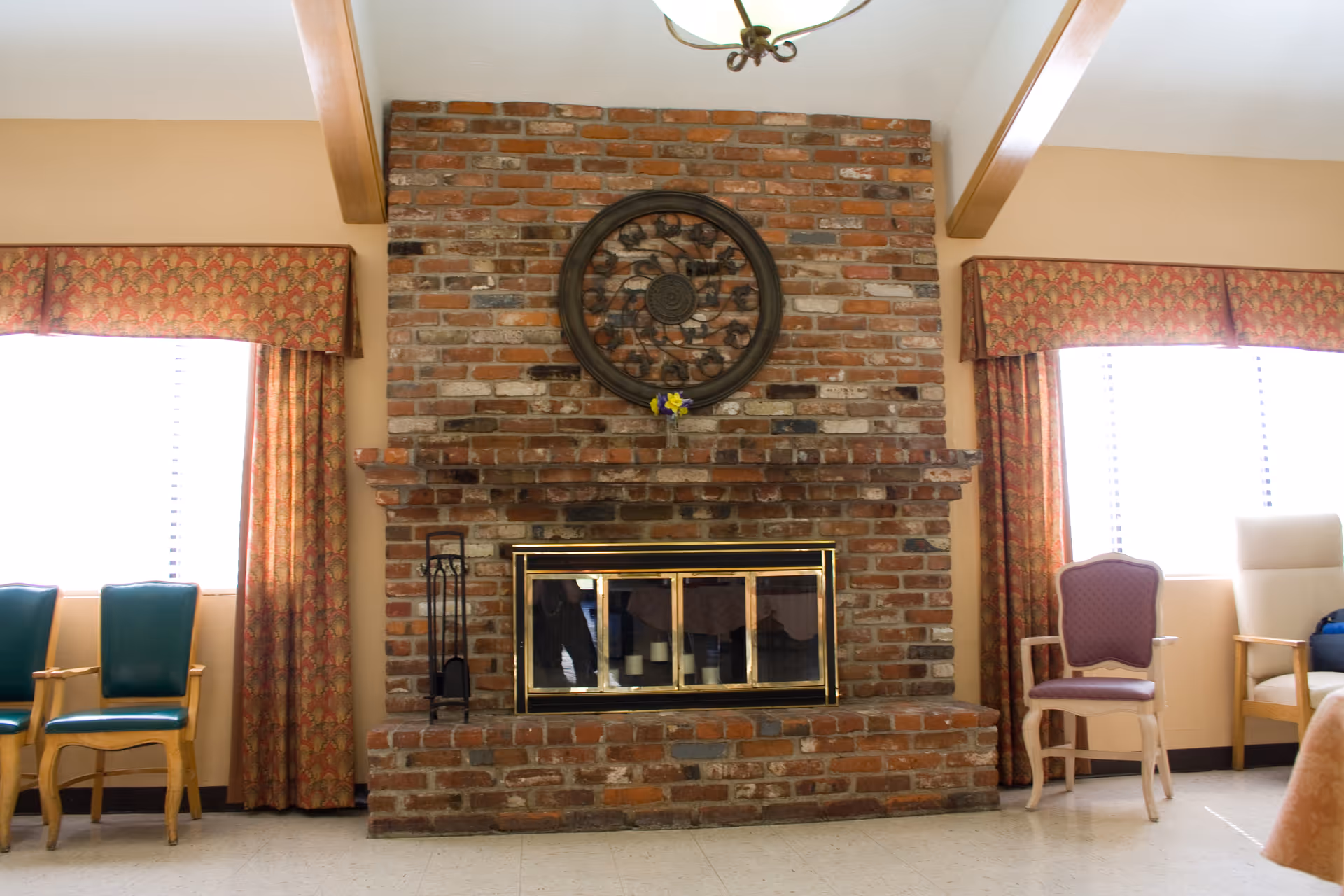 A cozy room with a large brick fireplace in the center, featuring a decorative circular metal wall art above it. On either side of the fireplace are windows with patterned curtains. There are several chairs arranged along the walls, including green, purple, and beige upholstered chairs. The ceiling has wooden beams and a hanging light fixture.