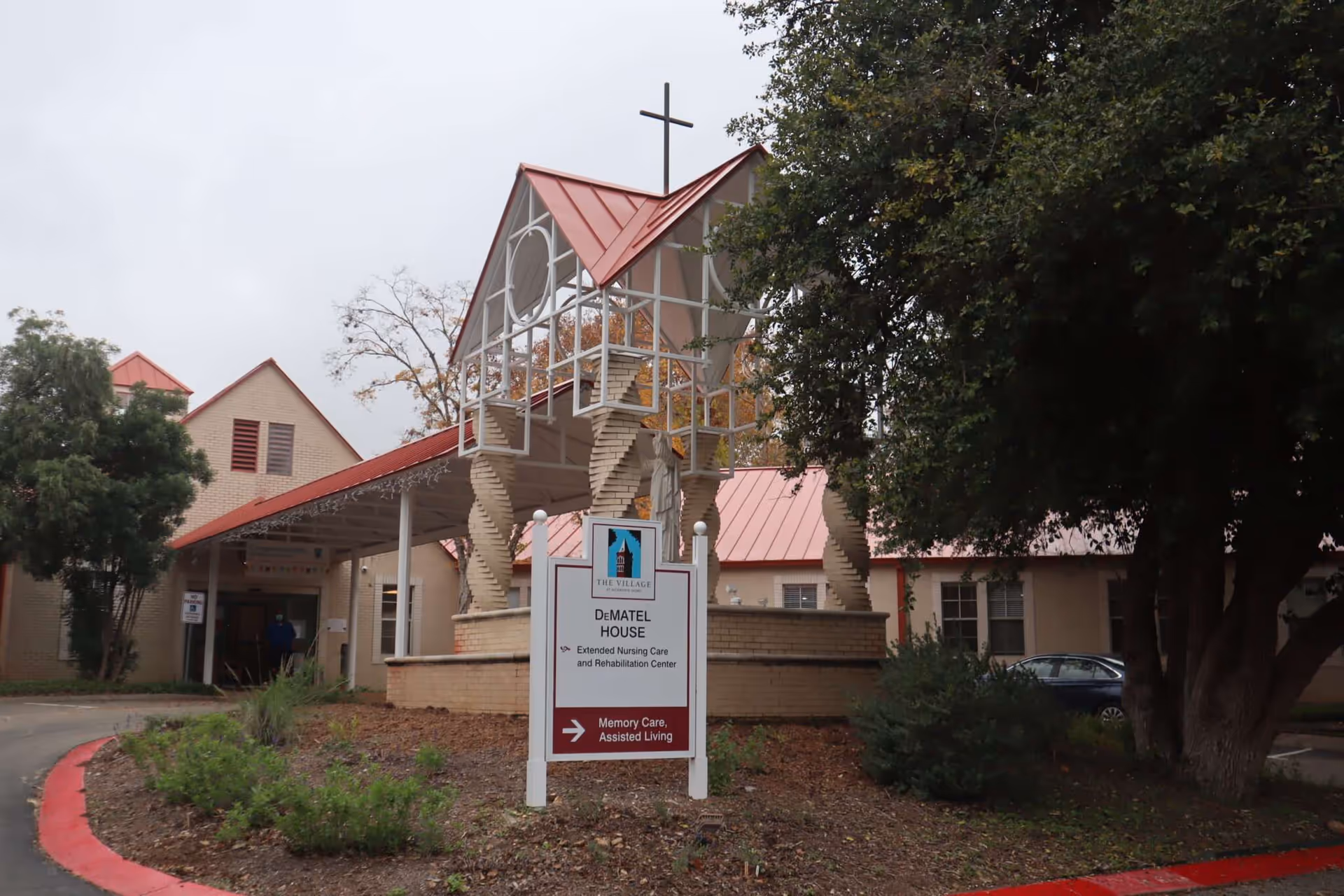 Exterior view of The Village at Incarnate Word's DeMatel House, an extended nursing care and rehabilitation center. The building has a red roof and beige brick walls, with a covered entrance and a sign indicating memory care and assisted living services. There is a large tree and some shrubs in the foreground.