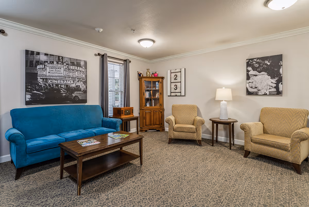 A cozy living room with a blue sofa, two beige armchairs, a wooden coffee table with magazines, a wooden cabinet, a side table with a lamp, and black and white wall art. The room has a patterned carpet and a window with gray curtains.