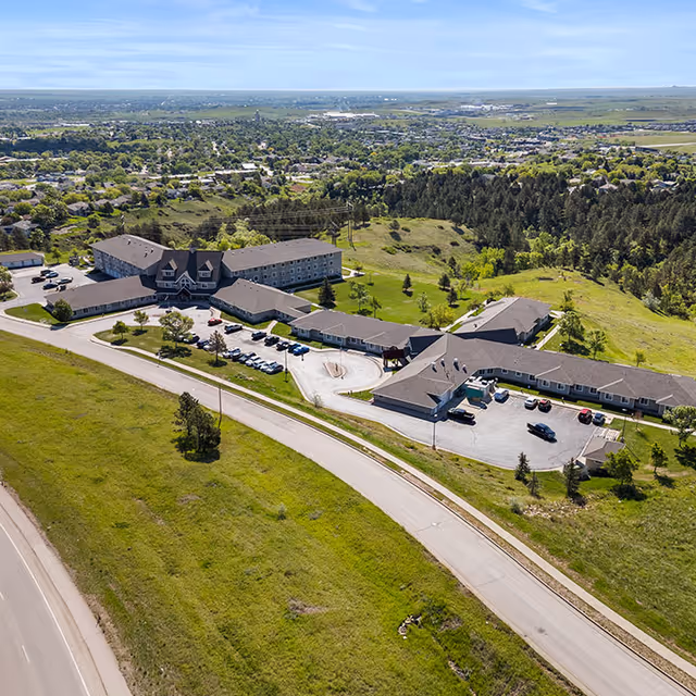 Aerial view of Good Samaritan Society - Echo Ridge - The Manor facility surrounded by green fields and trees, with a parking lot and multiple connected buildings under a clear blue sky.