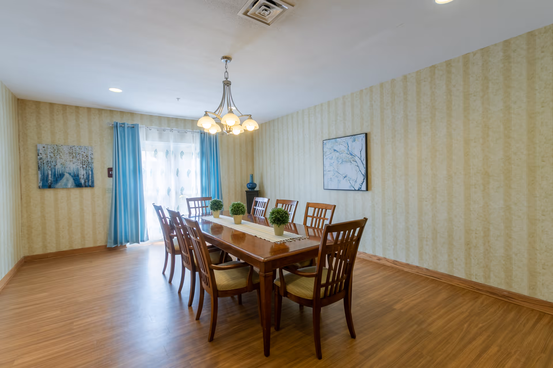 A dining room with a long wooden table surrounded by eight wooden chairs with cushioned seats. The table has a beige runner and three small potted plants as centerpieces. The room features light yellow patterned wallpaper, wood flooring, a chandelier with multiple lights hanging from the ceiling, and two paintings on the walls. Blue curtains frame a window with sheer white curtains allowing natural light to enter.