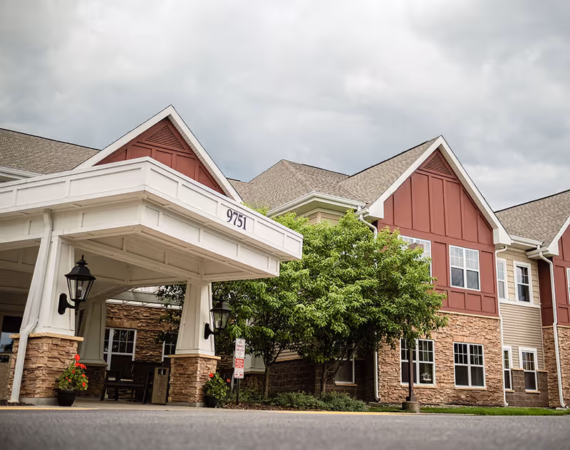 Front entrance of a senior living building with a covered porte-cochere labeled 9751, stone facade and red upper siding.