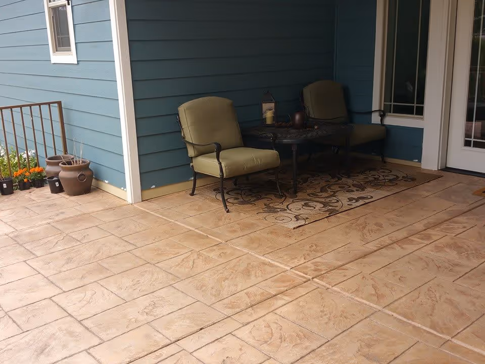 Outdoor patio area with two cushioned chairs and a small table on a decorative rug, next to a blue exterior wall with a window and a door. There are potted plants and flowers along the edge of the patio.