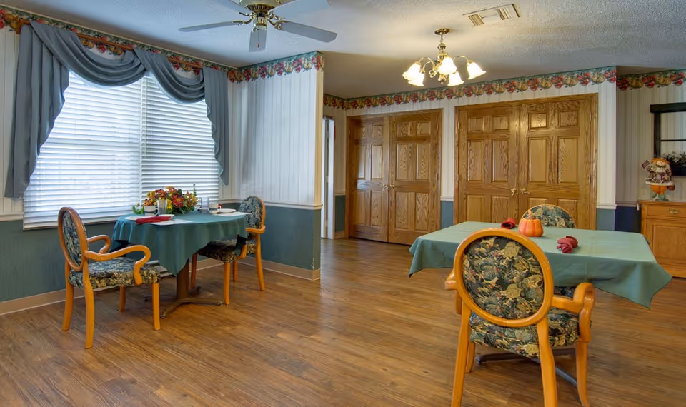 A dining room with two tables covered in green tablecloths, each surrounded by floral upholstered wooden chairs. One table has a centerpiece with flowers and the other has a small pumpkin decoration. The room has wood flooring, floral wallpaper border near the ceiling, and large windows with blue drapes and blinds. There are wooden double doors on one wall and a ceiling fan with lights above.