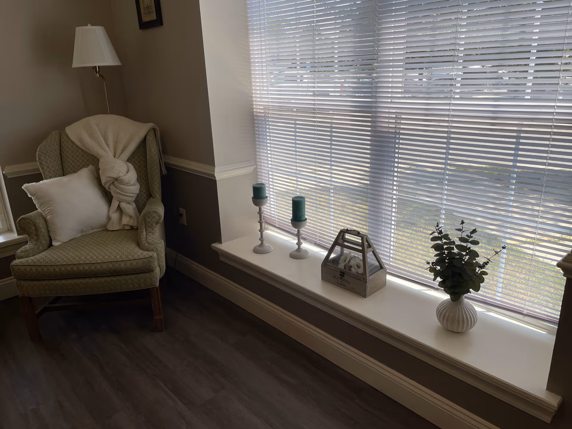 A cozy corner of a room featuring a green upholstered armchair with a white pillow and a white knitted blanket draped over the back. Next to the chair is a floor lamp with a white lampshade. A large window with white blinds lets in natural light, and the windowsill is decorated with two green candles on white holders, a small wooden box labeled 'FLOWERS & PLANTS' containing white flowers, and a white vase with green foliage.
