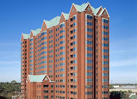 Tall multi-story red brick building with greenish peaked roofs under a clear blue sky, surrounded by some trees and distant structures.