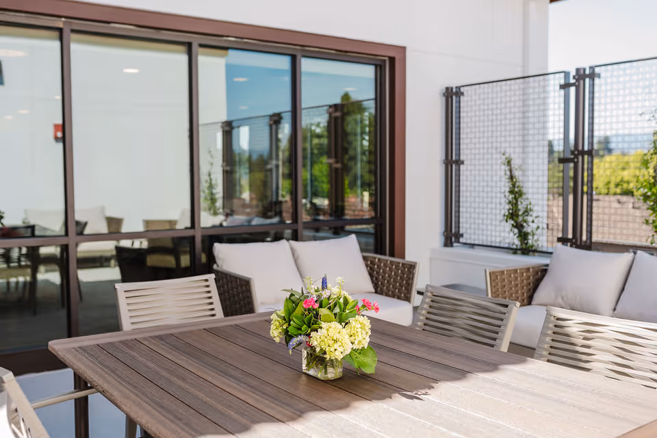 Patio dining area with a wooden table topped by a small vase of flowers, surrounded by chairs and cushioned sofas in front of sliding glass doors.