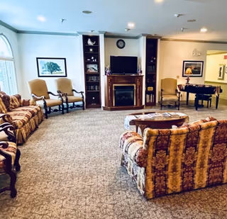 A cozy living room with patterned sofas and armchairs arranged around a wooden coffee table. There is a fireplace with a TV mounted above it, flanked by built-in shelves. The room has carpeted flooring, framed artwork on the walls, and a piano in the background near a hallway.