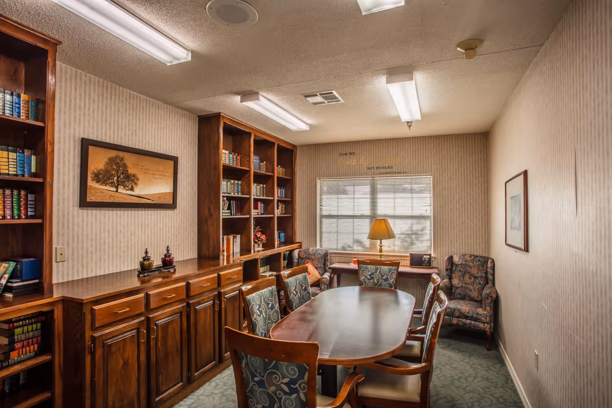 Small senior living common room with an oval meeting table, upholstered chairs, built-in bookshelves and a window with a lamp.