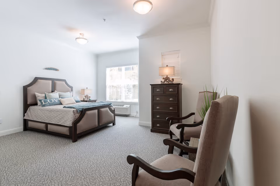 A bright and spacious bedroom with a large upholstered bed adorned with pillows and a blanket. There is a window with blinds letting in natural light, a dark wooden chest of drawers with a lamp on top, and two upholstered armchairs with wooden arms. The room has light-colored walls and carpeted flooring.