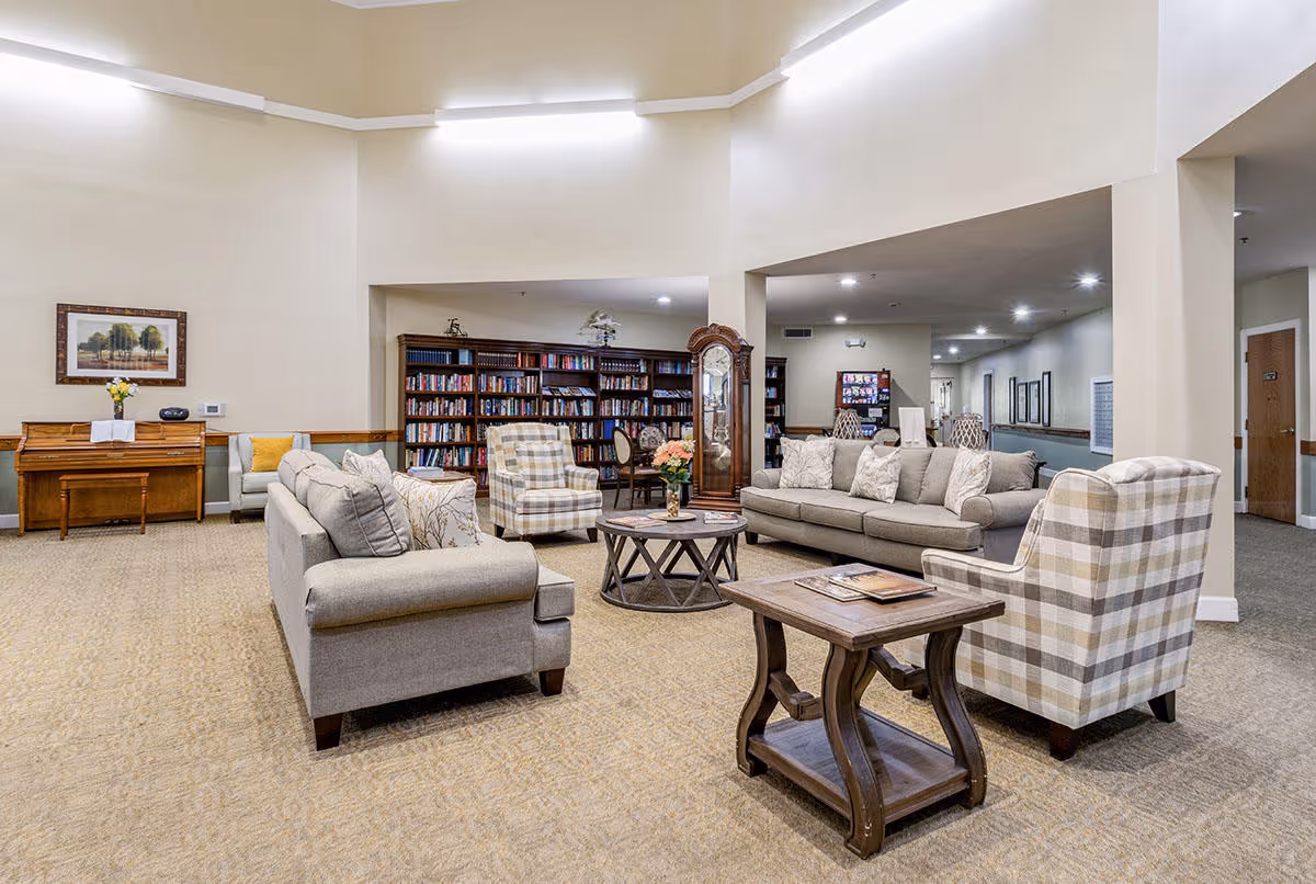 A spacious and well-lit living room area in a senior living facility with beige walls and carpet. The room features two gray sofas, two plaid armchairs, a wooden coffee table with flowers, a side table with magazines, a grandfather clock, and a bookshelf filled with books. There is also a piano against one wall with a framed picture above it.