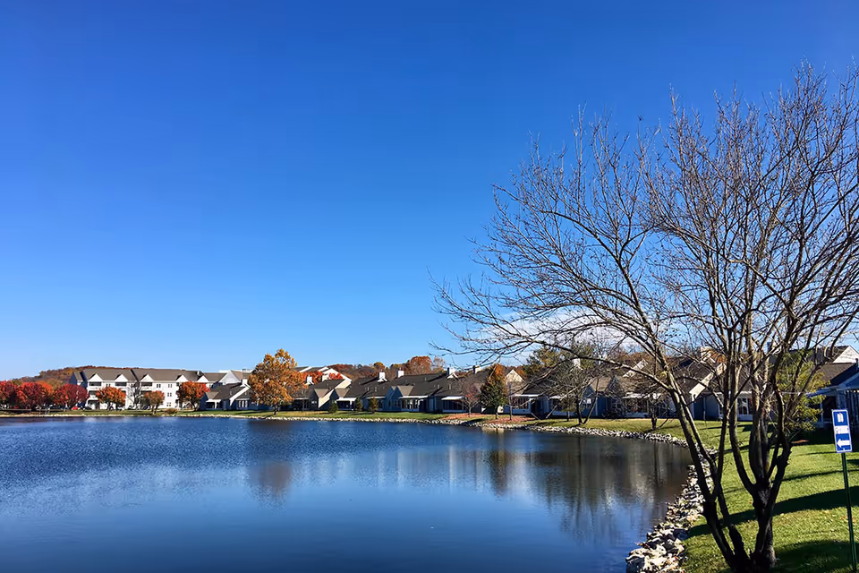 A serene view of a pond with clear blue water reflecting the sky and surrounding buildings. On the far side of the pond, there are multiple residential buildings with autumn-colored trees interspersed. In the foreground, there is a leafless tree on the right side and a grassy area with a signpost.