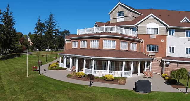 Front exterior of a multi-story senior living building with a wraparound porch, patio seating, flagpole, and landscaped lawn under a clear blue sky.
