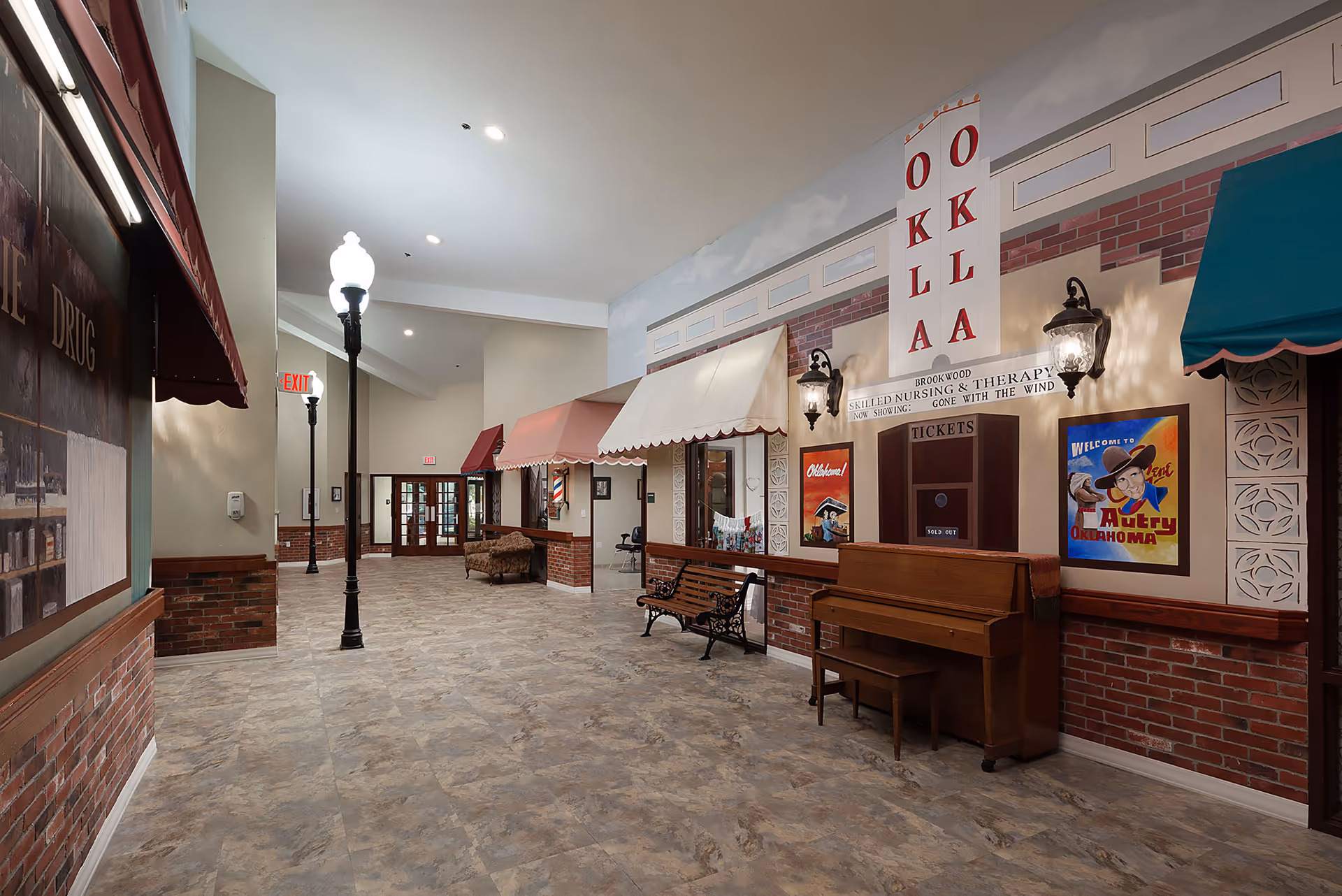 Interior hallway of a senior living facility styled like a small-town street with storefront facades, benches, a piano, and street lamps.