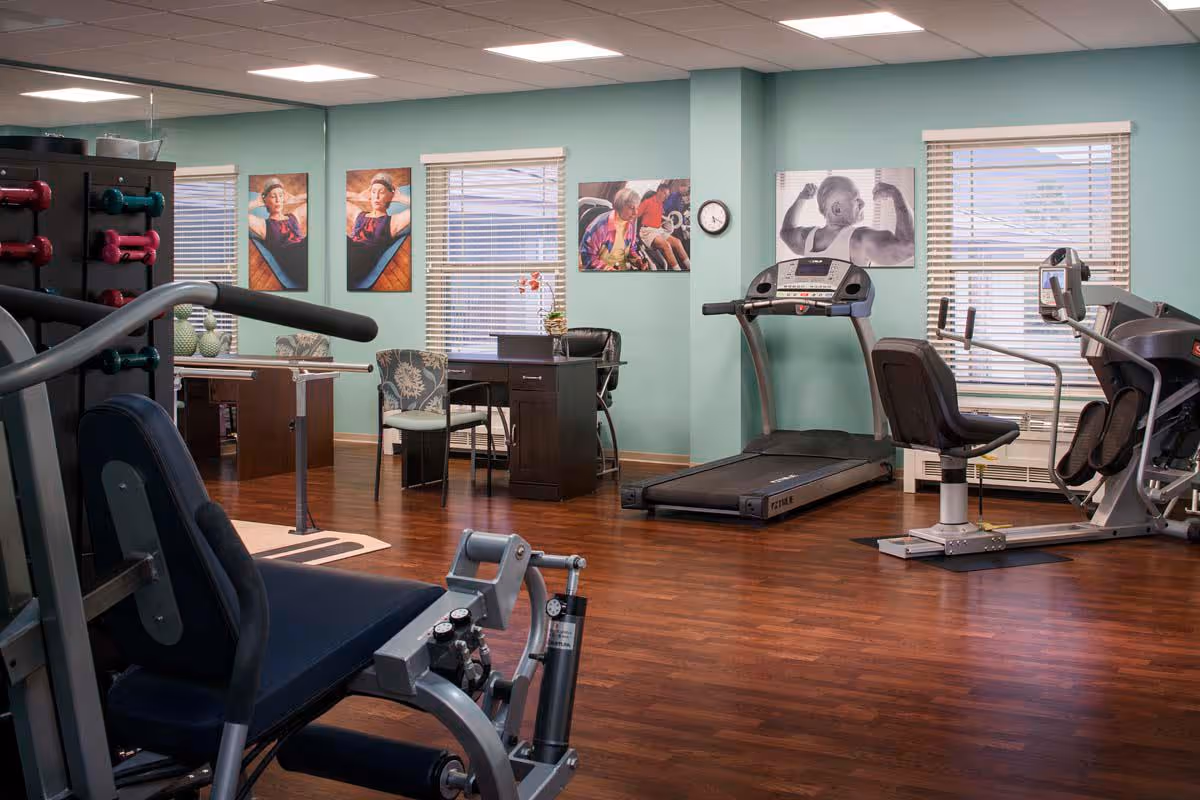 A senior living facility exercise room with various gym equipment including a treadmill, stationary bike, weight machine, and dumbbells. The room has wooden flooring, light blue walls, three windows with blinds, and several framed pictures on the walls. There is also a desk with two chairs near the windows.
