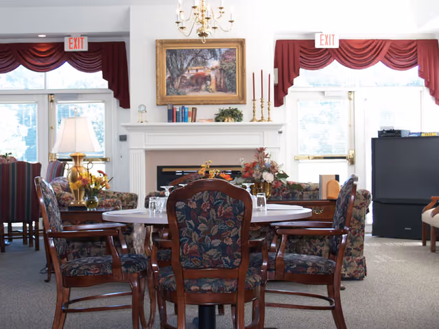 A cozy living room area with a round table surrounded by four floral upholstered wooden chairs in the foreground. Behind the table is a white fireplace mantel decorated with books, candles, and a plant, with a framed painting hanging above it. To the left, there is a table lamp and more floral upholstered chairs. The room has large windows with red valances and two exit signs above the doors. A large television is visible on the right side of the room.