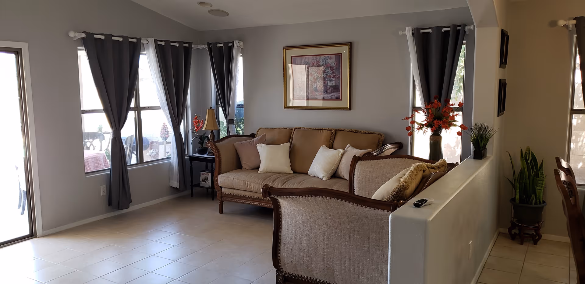 Sunlit living room with a beige sofa and throw pillows, gray curtains at multiple windows, wall art, and tiled floor.