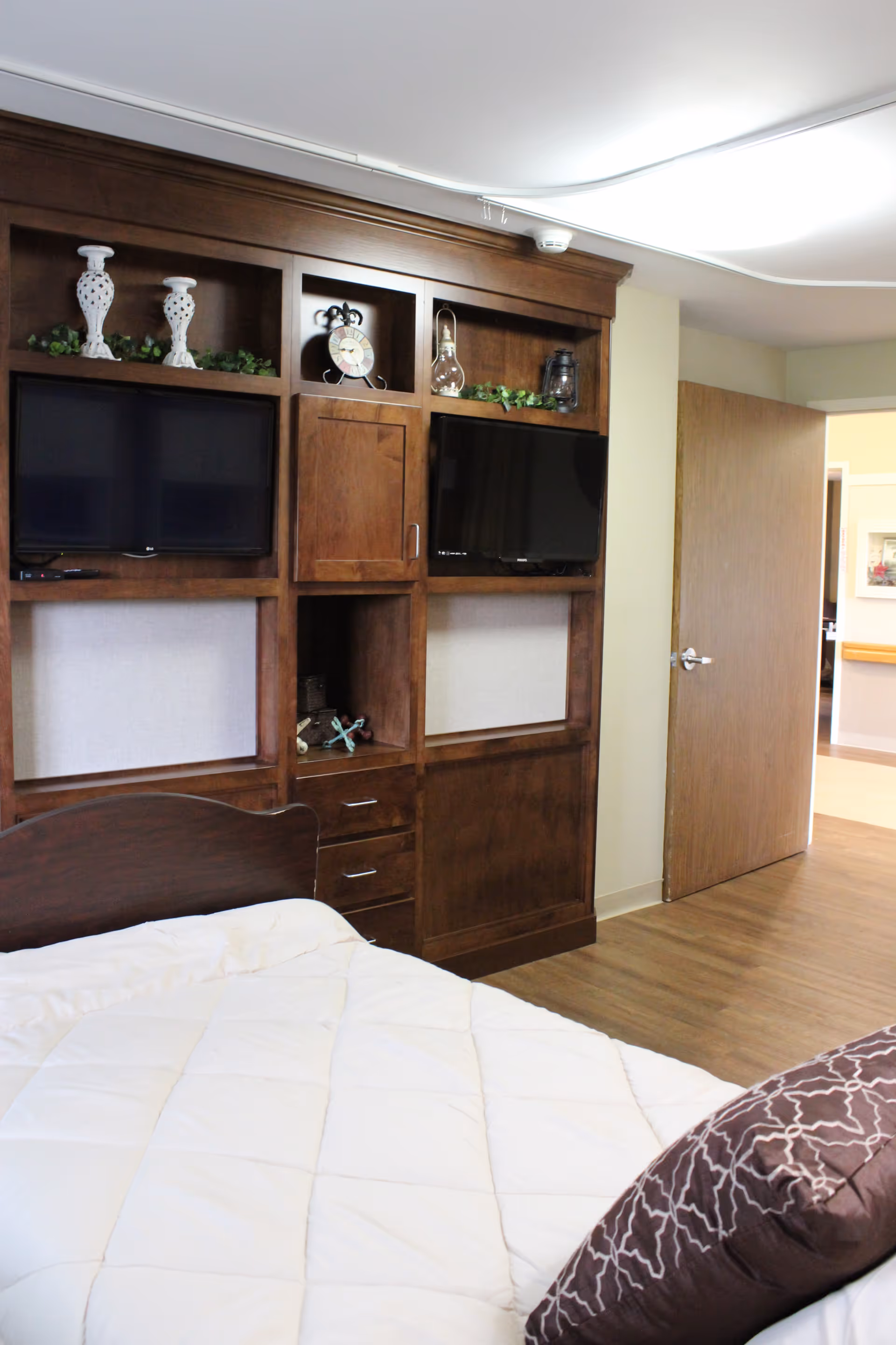 A bedroom in a senior living facility featuring a bed with a white comforter and a brown pillow. There is a large wooden entertainment unit with two flat-screen TVs, decorative items including vases, a clock, and greenery. The room has wooden flooring and an open door leading to a hallway.