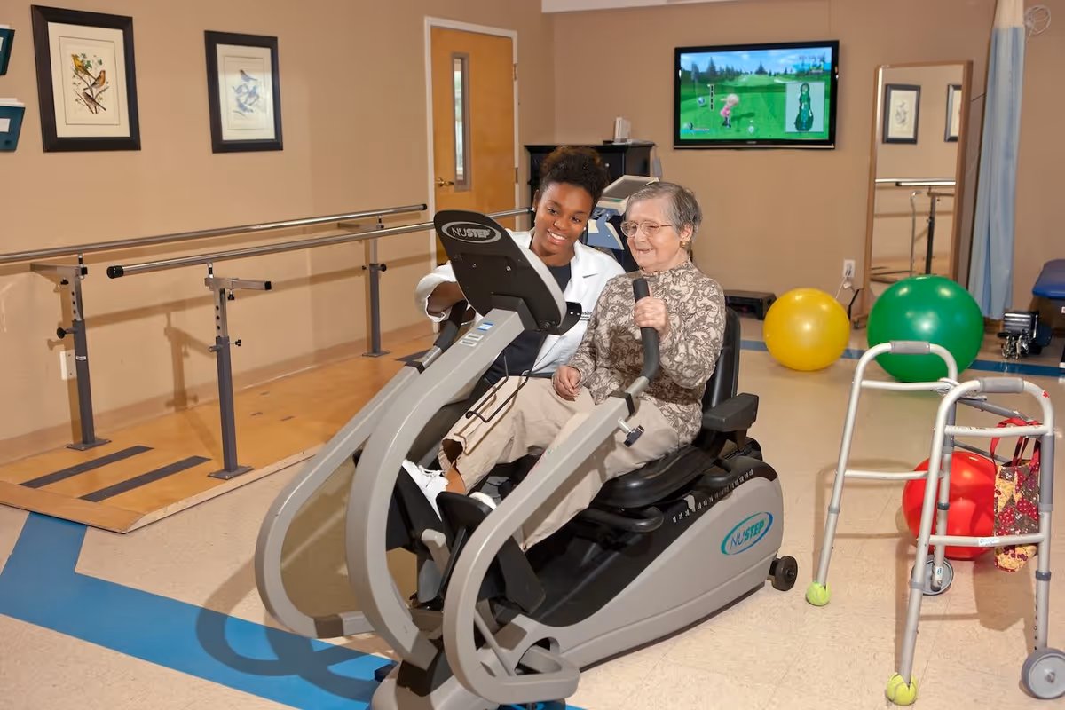An elderly woman is seated on a NuStep exercise machine in a rehabilitation room, assisted by a healthcare professional in a white coat. The room has handrails along one wall, colorful exercise balls, a walker, and a television mounted on the wall displaying a golf video game.