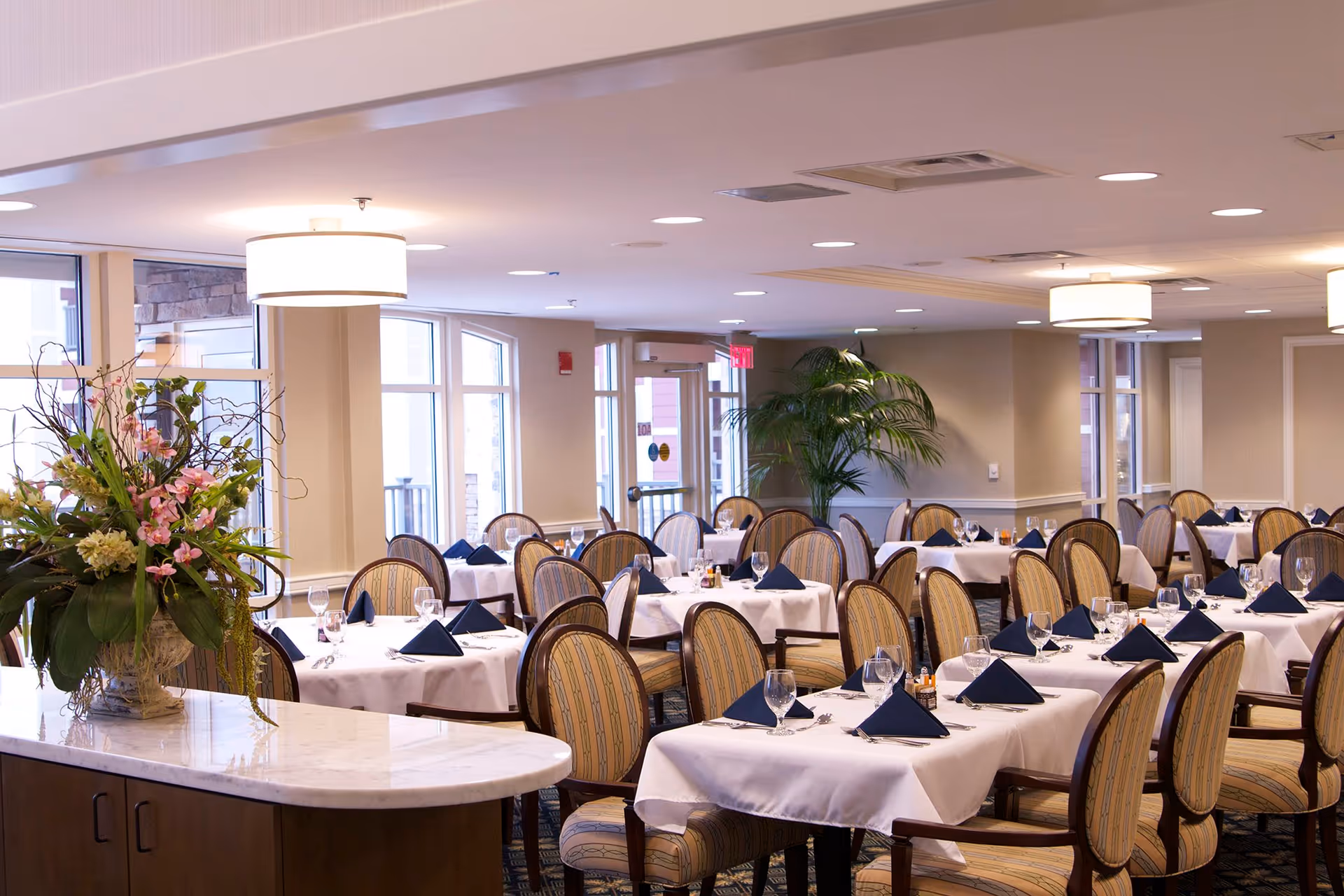A spacious dining room with multiple tables covered in white tablecloths, each set with navy blue folded napkins, wine glasses, and silverware. The room features upholstered chairs with striped fabric, large windows allowing natural light, ceiling lights, and a large floral arrangement on a marble countertop in the foreground.