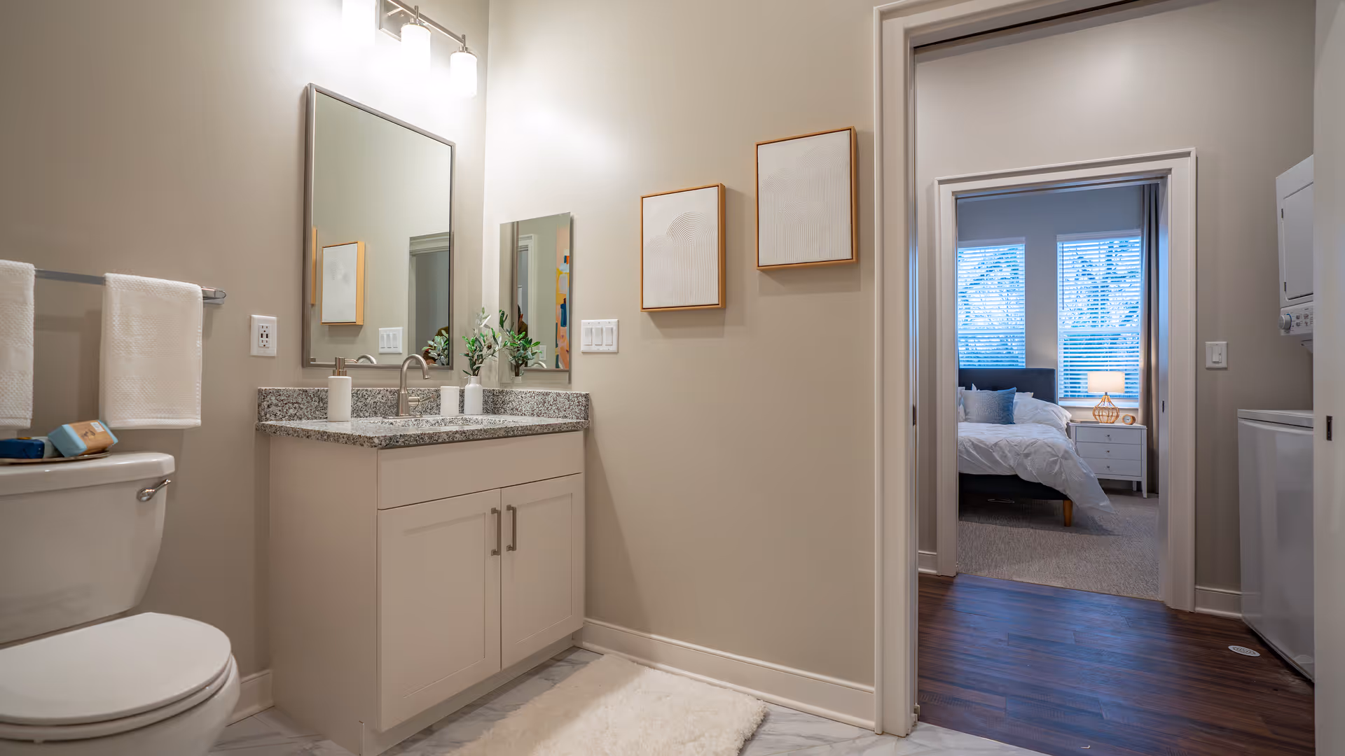 Modern bathroom with a granite-topped vanity, mirror and toilet, towels on a rack, and a view through a doorway into a bedroom.
