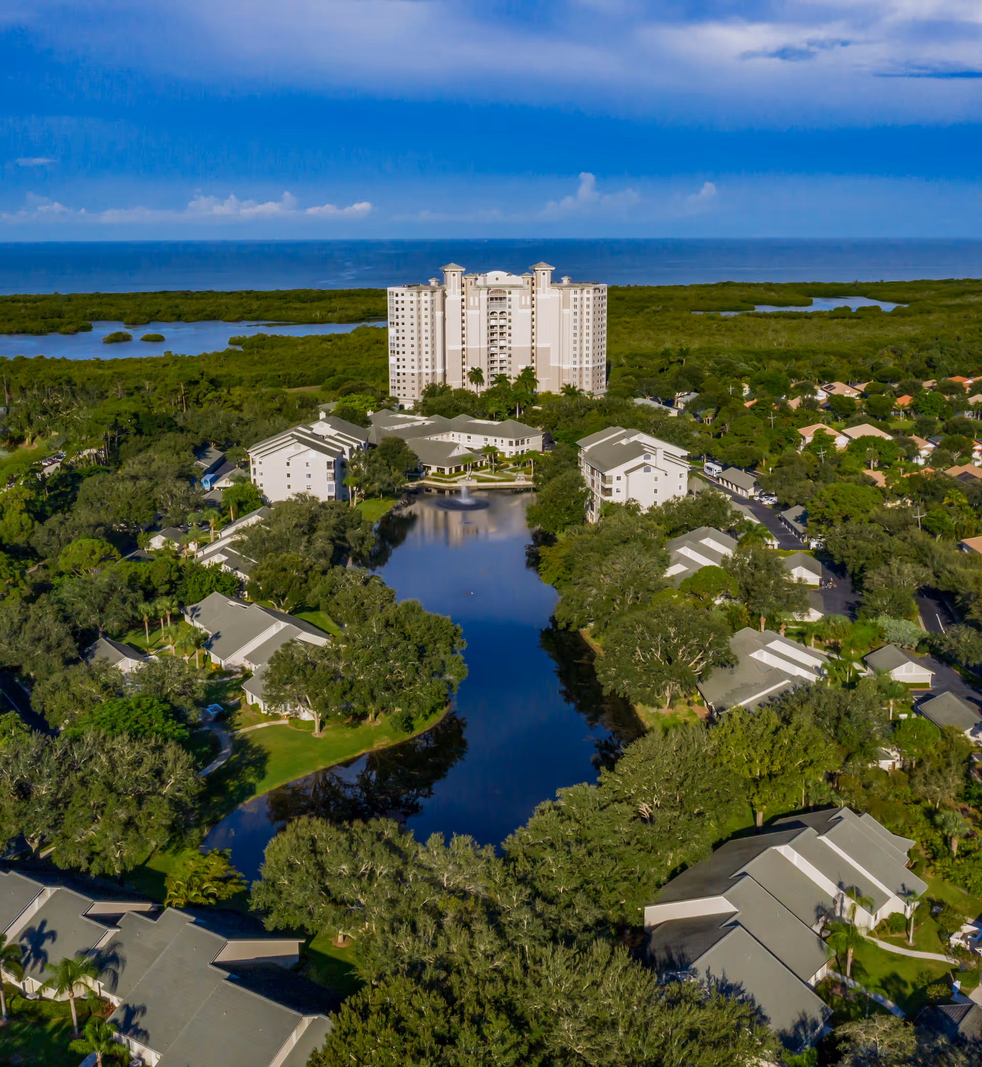 Aerial view of Arbor Trace senior living facility showing multiple residential buildings surrounding a central pond with a fountain, lush green trees, and a large multi-story building in the background with a body of water and blue sky beyond.