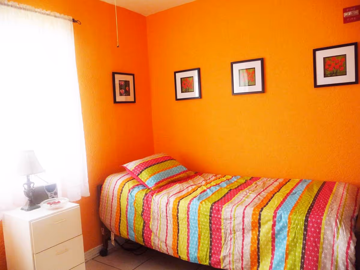 Small bedroom with bright orange walls, a single bed with colorful striped bedding, a nightstand with a lamp, and framed pictures above the bed.