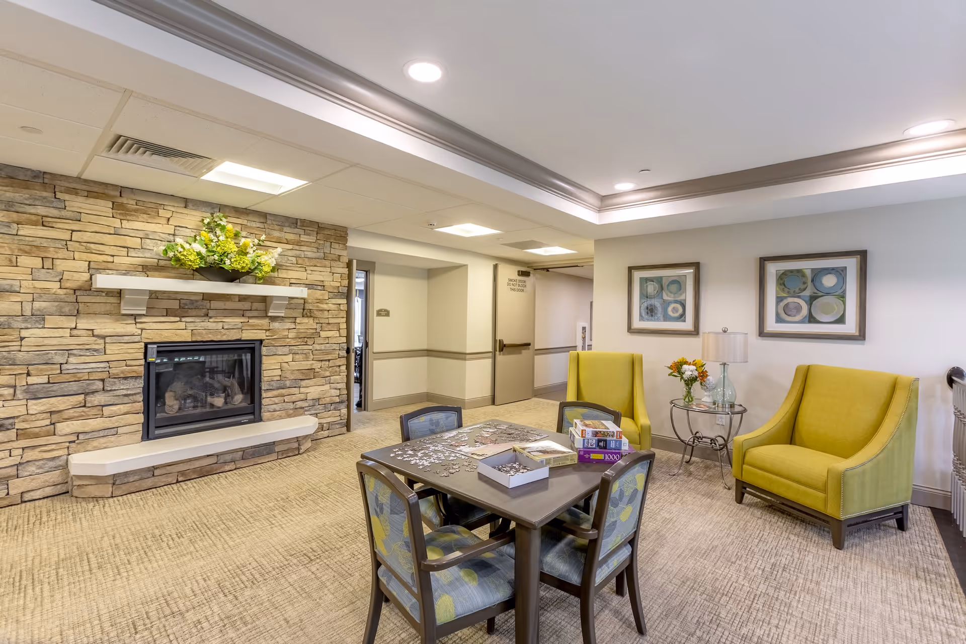 A cozy common area in a senior living facility featuring a stone fireplace with a flower arrangement on the mantel, a table with chairs around it holding puzzle pieces and board games, two yellow armchairs, a small glass side table with a lamp and flowers, and framed abstract artwork on the walls.