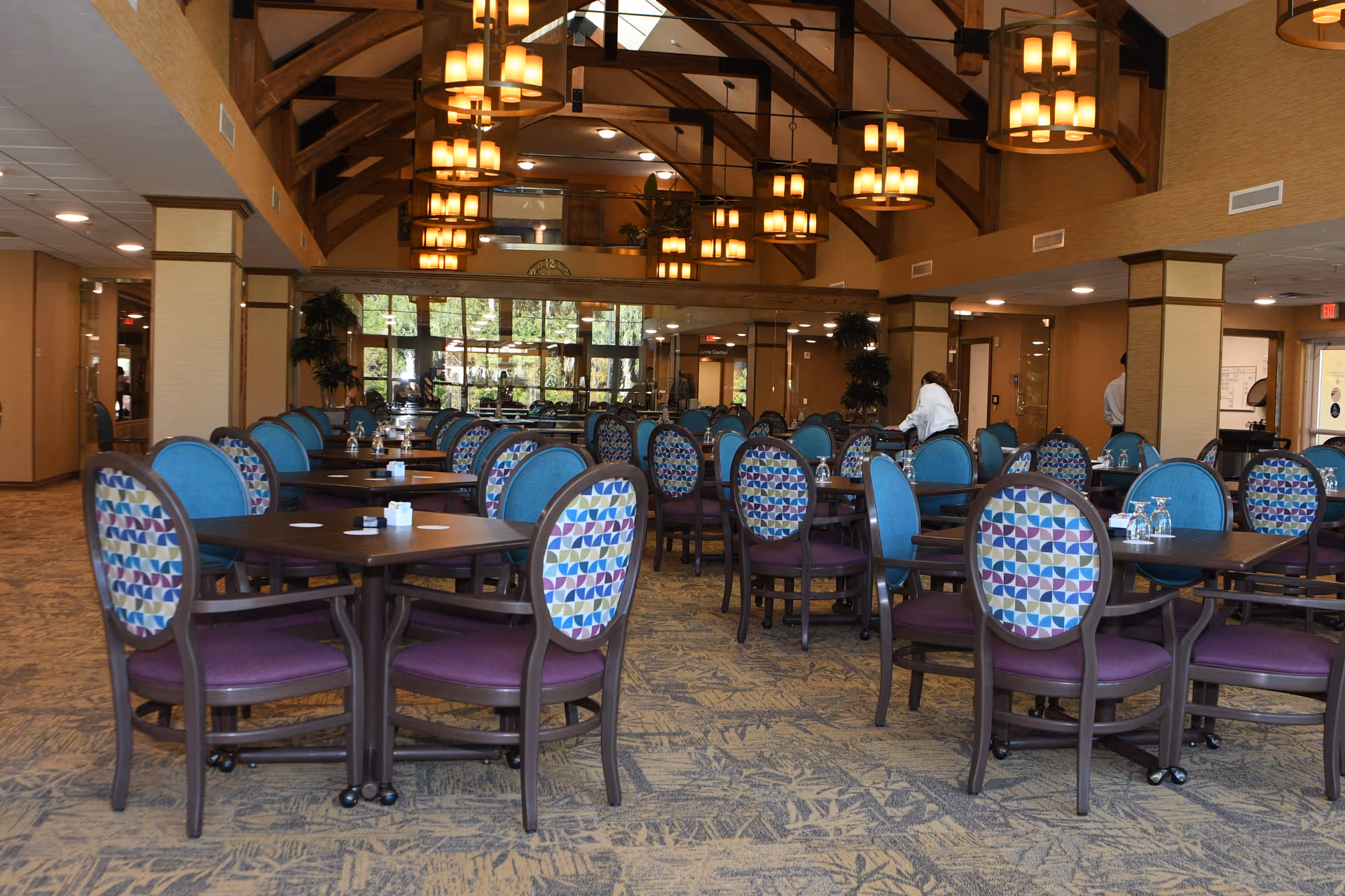 A spacious dining room in Palomino Gardens Retirement Community with multiple tables and chairs arranged neatly. The chairs have colorful patterned backs and purple seats. The room features high vaulted ceilings with wooden beams and several hanging light fixtures. Large windows at the far end allow natural light to enter, and a few people are seen in the background near the walls.