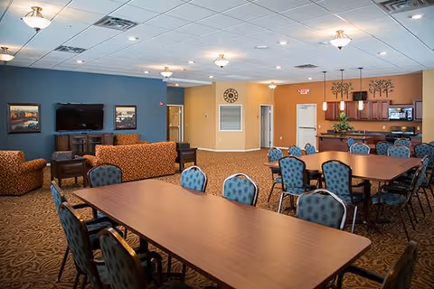 A spacious senior living common area with multiple tables and chairs arranged for dining or activities. The room features a seating area with patterned sofas and armchairs facing a flat-screen TV mounted on a blue accent wall. The back wall includes a kitchen area with wooden cabinets, a microwave, and pendant lighting. The walls are painted in warm tones of blue, beige, and orange, and the ceiling has recessed lighting fixtures.