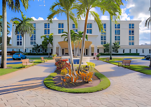 Front exterior view of a multi-story assisted living facility with a paved driveway, landscaped garden with palm trees, benches, and a clear blue sky.