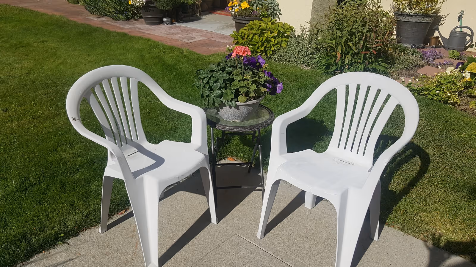 Two white plastic lawn chairs with a small round table and potted flowers on a patio next to a grassy garden.