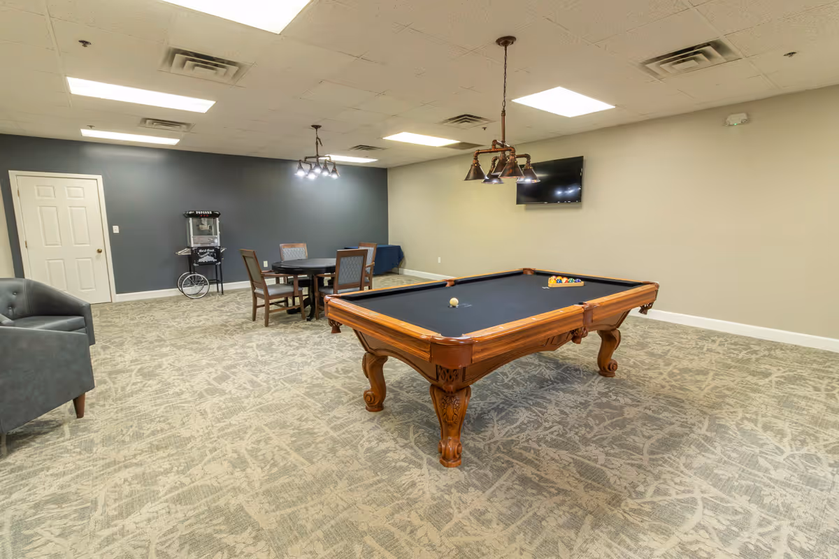 A spacious recreational room with a pool table in the center, a round table with four chairs in the background, a popcorn machine against a dark gray wall, a mounted flat-screen TV on a beige wall, and a gray armchair on the left side. The room has patterned carpet flooring and ceiling lights.