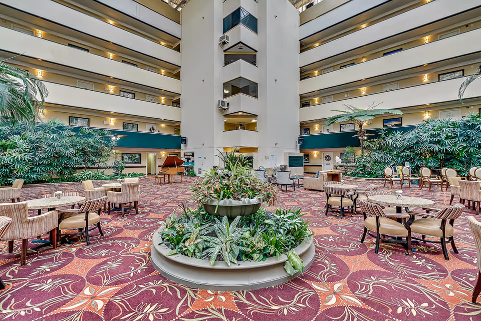Spacious indoor common area with patterned carpet, multiple seating arrangements including round tables with chairs and lounge chairs, a central planter with green plants, and a grand piano near the back. The area is surrounded by multiple floors with balconies and doors, and large green plants line the walls.