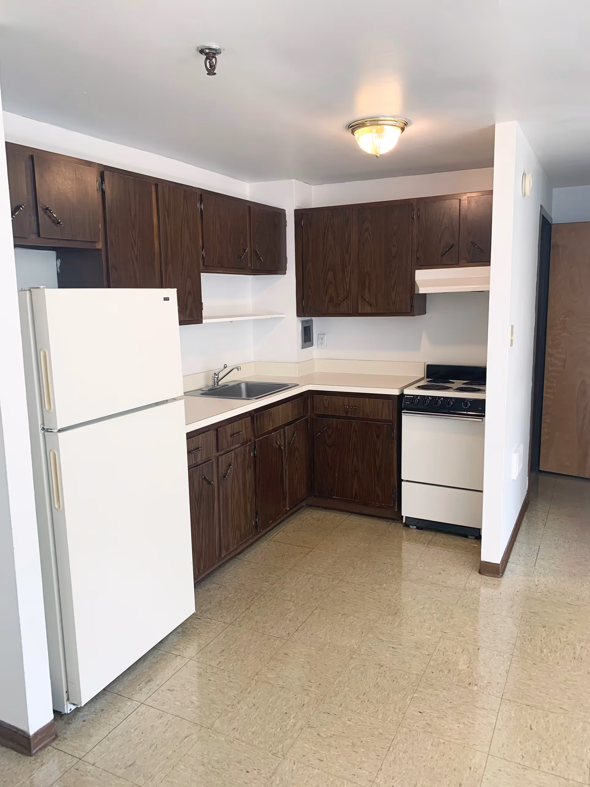 Small kitchenette with a white refrigerator, sink, stove, and dark wood cabinets in a compact apartment space.