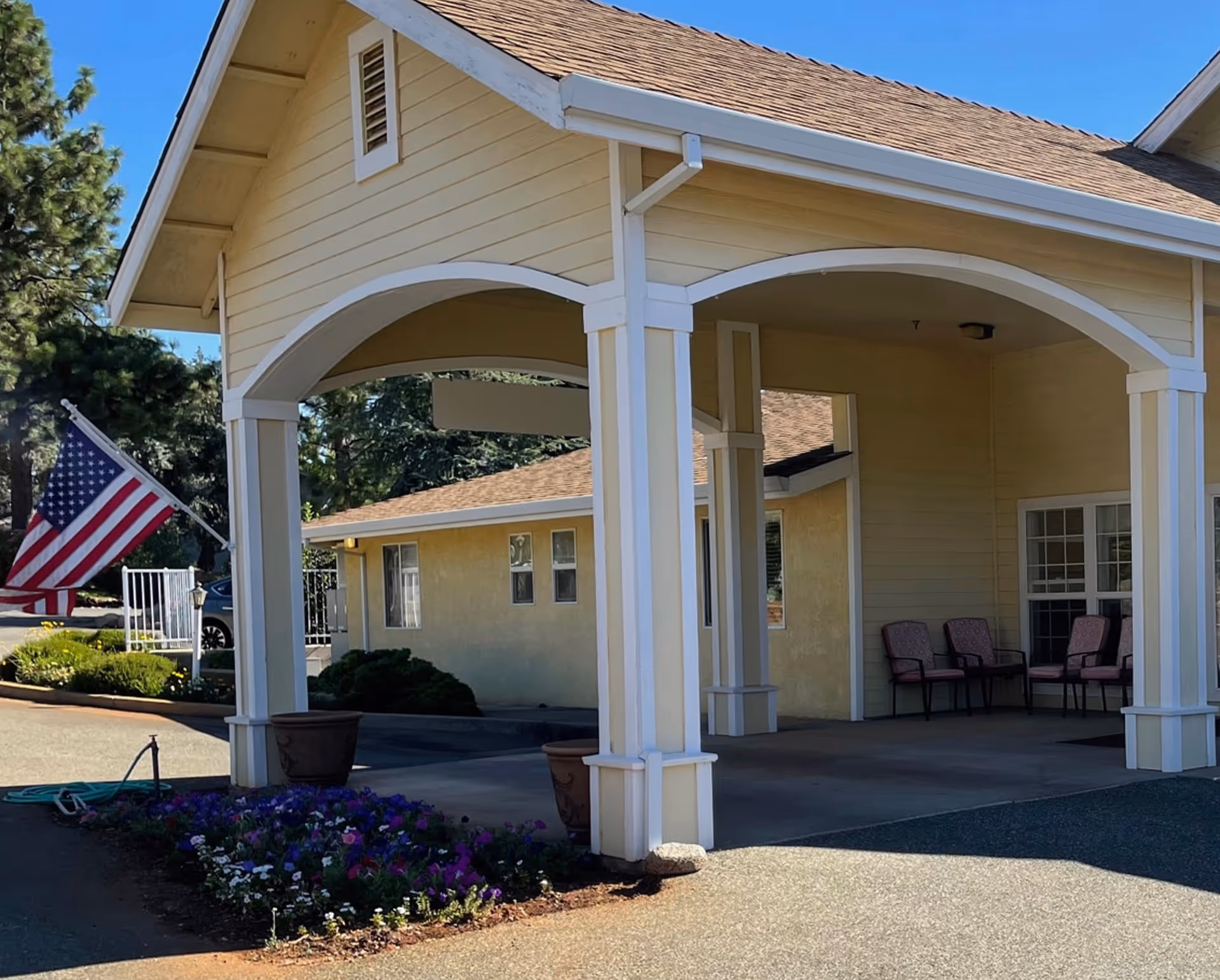 Covered entrance/portico of a yellow building with an American flag, potted plants, chairs, and a flowerbed.