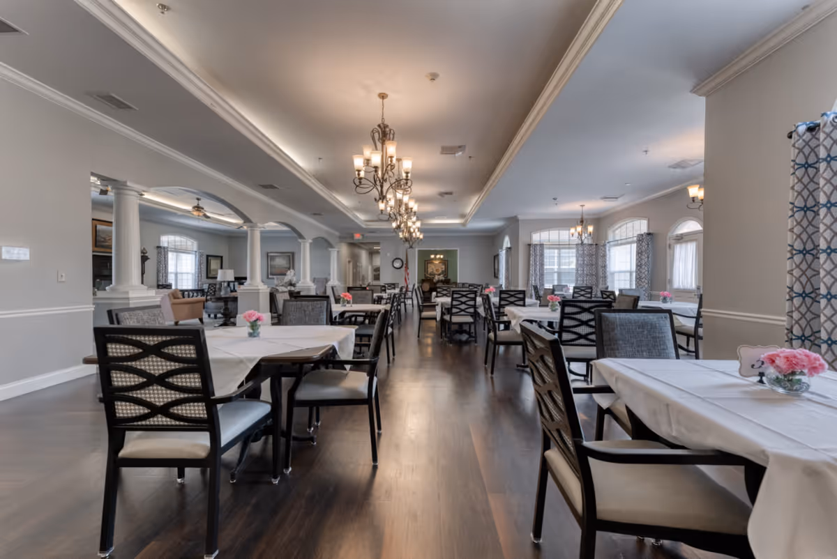 A spacious dining room with multiple tables covered in white tablecloths, each adorned with small pink flower arrangements. The room features dark wooden floors, elegant chandeliers hanging from a tray ceiling, and large windows with patterned curtains allowing natural light to fill the space. There are several chairs around each table, and the room has a calm, inviting atmosphere.