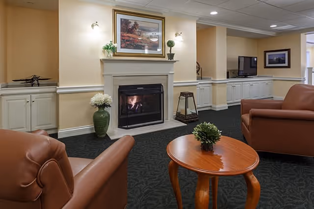 A cozy living room area with two brown leather armchairs facing a fireplace with a fire burning. Above the fireplace is a framed painting and two wall sconces. There are built-in white cabinets on either side of the fireplace, a green vase with white flowers to the left, and a small wooden table with a green plant in the center. A flat-screen TV is mounted on the wall in the background.