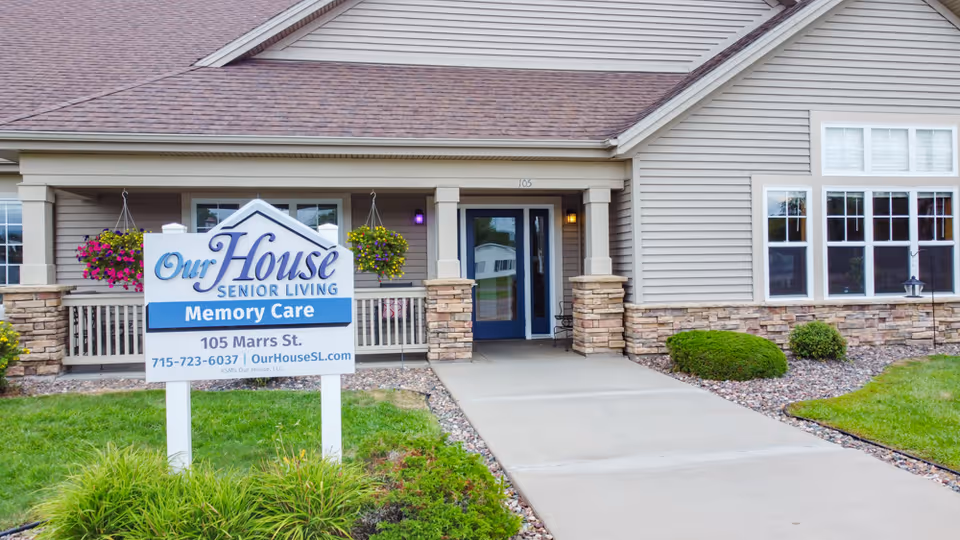 Exterior front entrance of Our House Senior Living Memory Care facility at 105 Marrs St., featuring a sign with contact information, a concrete walkway, hanging flower baskets, and a beige building with stone accents and windows.