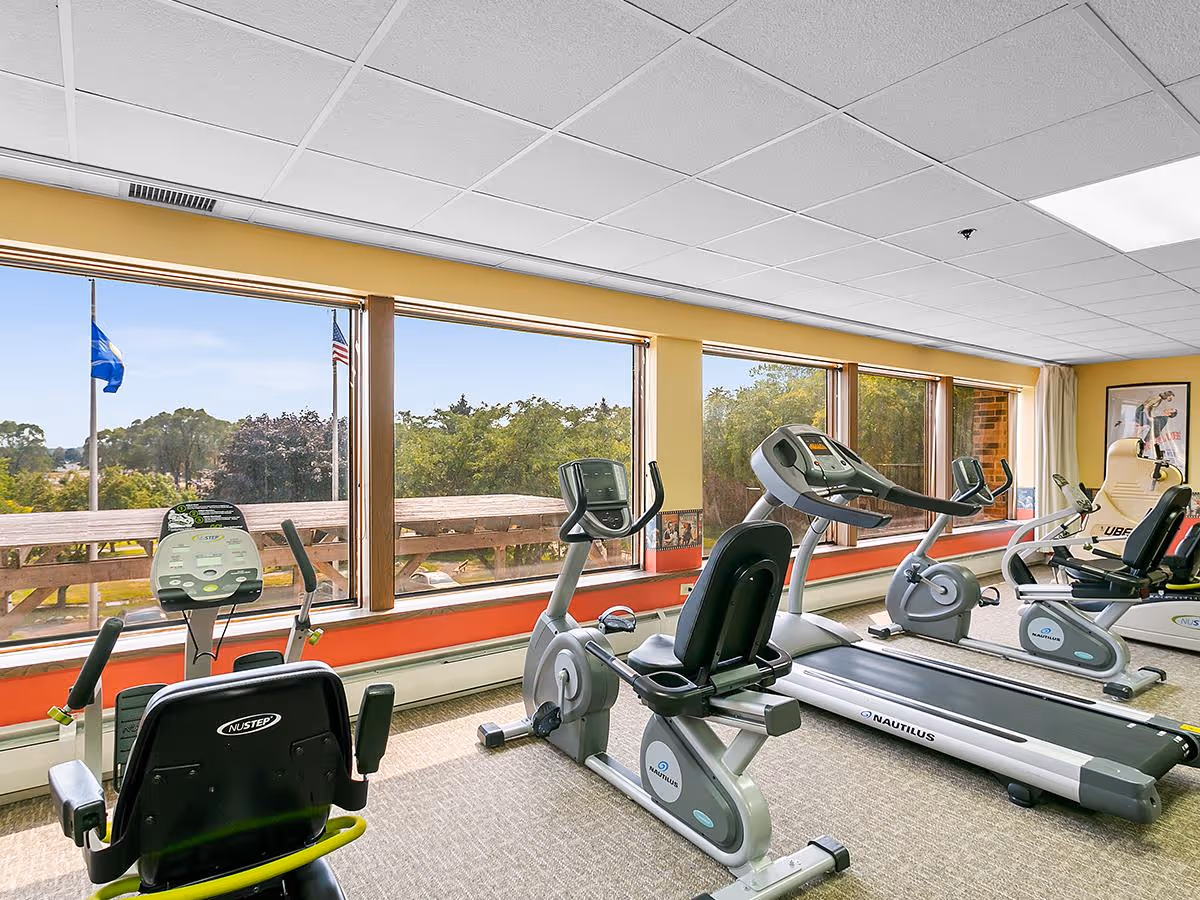 A fitness room with exercise equipment including stationary bikes and a treadmill, large windows showing an outdoor view with flags and trees, and a bright, spacious interior with yellow walls and carpeted floor.