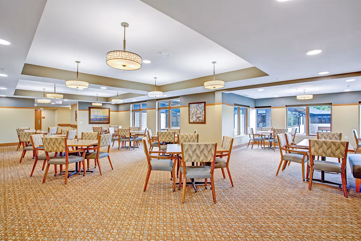 Spacious communal dining room with multiple tables and chairs, patterned carpet, large windows and pendant ceiling lights.