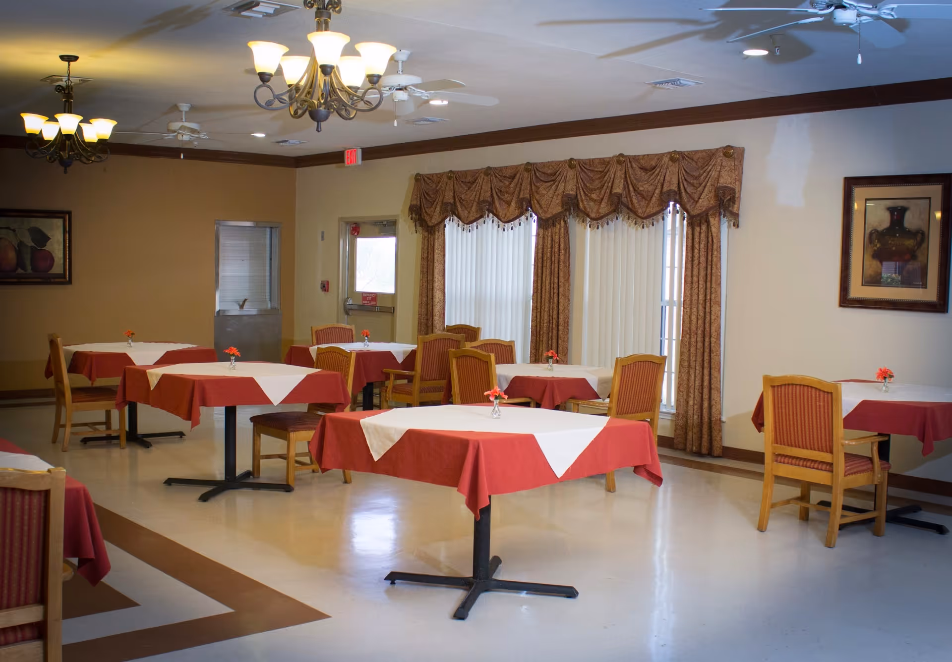 A dining room with several tables covered with red tablecloths and white overlays, each table having a small vase with a red flower. The room has beige walls, framed artwork, large windows with vertical blinds and decorative curtains, ceiling fans, and chandeliers providing light.