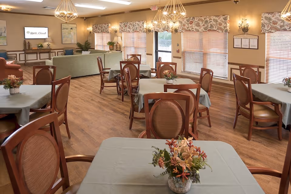 Communal dining and lounge area with multiple tables and chairs, floral centerpieces, and a seating area with sofas and a TV showing the Apple Creek logo.