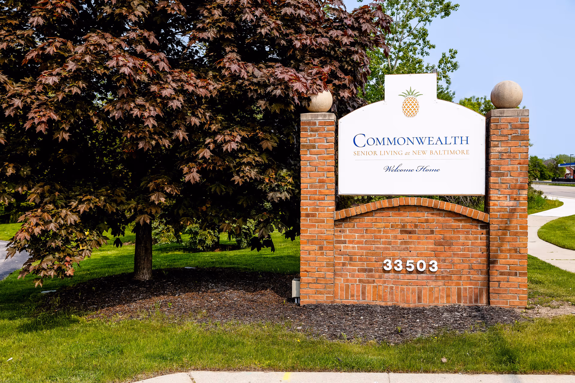 A brick signpost with a white sign displaying the text 'Commonwealth Senior Living at New Baltimore Welcome Home' and a pineapple logo above the text. The signpost is surrounded by green grass and a tree with dark red leaves on the left side. A sidewalk curves around the sign on the right side under a clear blue sky.